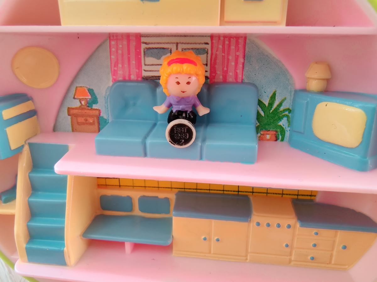 A miniature sized Polly Pocket plastic doll sitting on a blue couch inside a doll house.